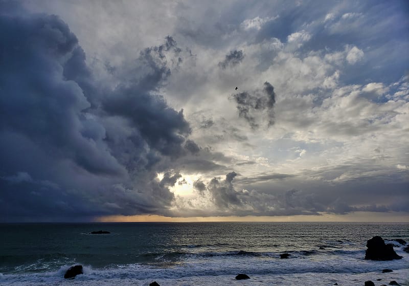 Towering dark storm clouds moving in over the Pacific surf with rain, while sunlight shines through a hole in the sky.