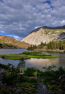 Alpine lake under stormy skies reflecting white granite, with lush green island in the center and shifting light, in the Sierra Nevada mountains near Yosemite.